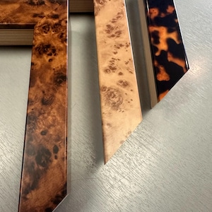 May include: Three wooden picture frame corner samples featuring distinct finishes. One displays a dark brown burl wood grain, another a lighter tan burl wood pattern, and the third a glossy black and orange tortoiseshell pattern.