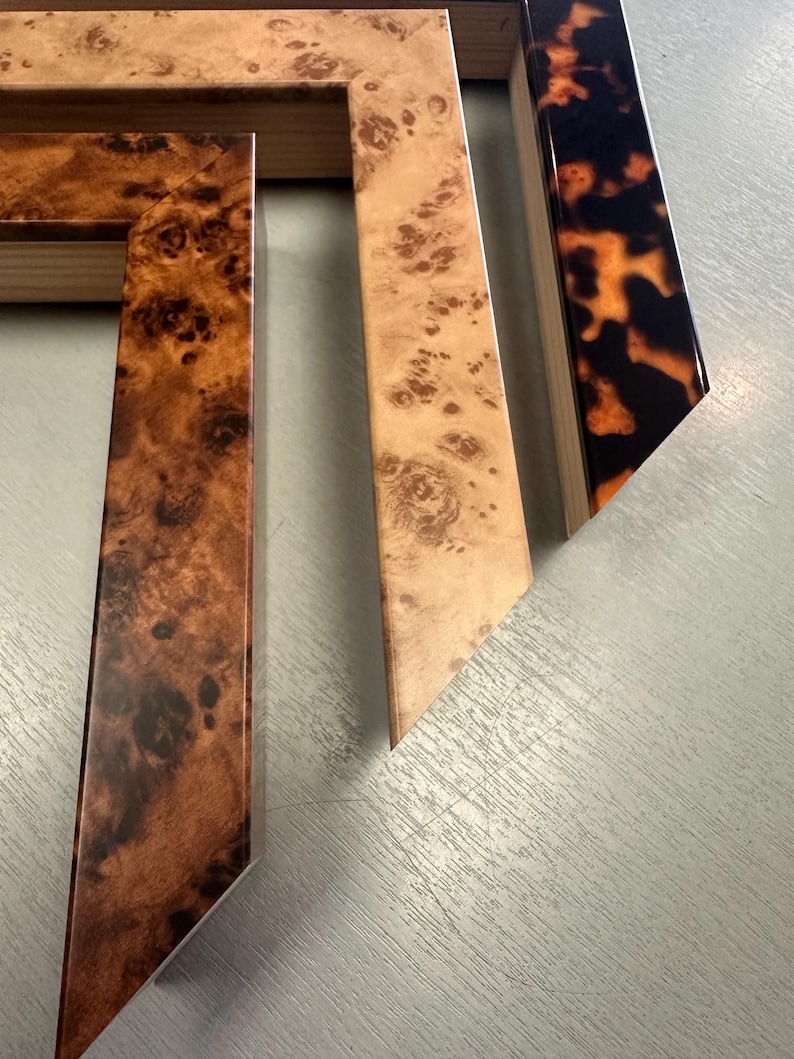 May include: Three wooden picture frame corner samples featuring distinct finishes. One displays a dark brown burl wood grain, another a lighter tan burl wood pattern, and the third a glossy black and orange tortoiseshell pattern.