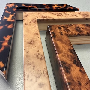 May include: Three L-shaped picture frame corners arranged in a stack. The top frame features a dark brown and orange tortoiseshell pattern. The middle frame has a light tan burl wood grain finish. The bottom frame displays a dark brown burl wood.