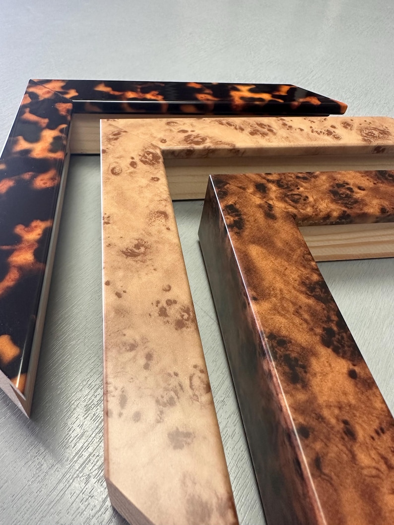 May include: Three L-shaped picture frame corners arranged in a stack. The top frame features a dark brown and orange tortoiseshell pattern. The middle frame has a light tan burl wood grain finish. The bottom frame displays a dark brown burl wood.