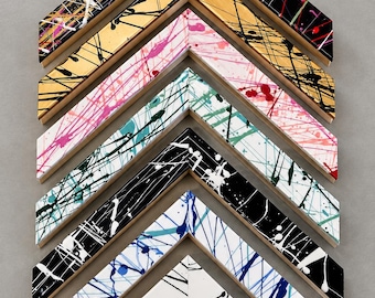May include: A collection of six V-shaped picture frame corner samples arranged in a vertical stack. Each frame features a unique base colour including black, gold, pink, white, and blue, all decorated with vibrant, multi-coloured paint splatter patterns.