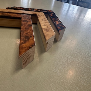 May include: Three L-shaped corner sections of picture frames arranged in a row. The first frame corner features a dark brown burl wood grain pattern, the second a lighter tan burl wood grain, and the third a dark tortoiseshell pattern finish.