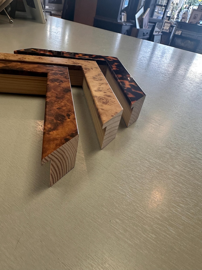 May include: Three L-shaped corner sections of picture frames arranged in a row. The first frame corner features a dark brown burl wood grain pattern, the second a lighter tan burl wood grain, and the third a dark tortoiseshell pattern finish.