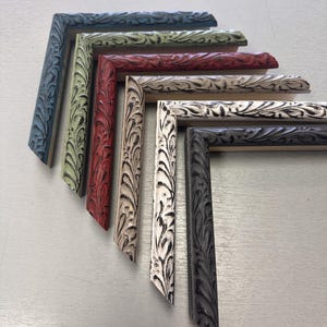 Puede incluir: Una colección de seis muestras de esquinas de marcos de fotos en forma de L dispuestas en abanico. Cada marco presenta un patrón de relieve ornamentado con forma de hoja con un acabado desgastado en azul, verde claro, rojo, beige, blanco y gris oscuro.