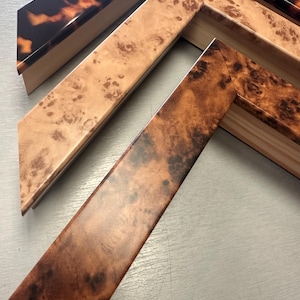 May include: Three L-shaped picture frame corner samples featuring distinct finishes. The top piece has a glossy tortoiseshell pattern, the middle piece displays a light tan burl wood grain, and the bottom piece shows a darker brown burl wood finish.