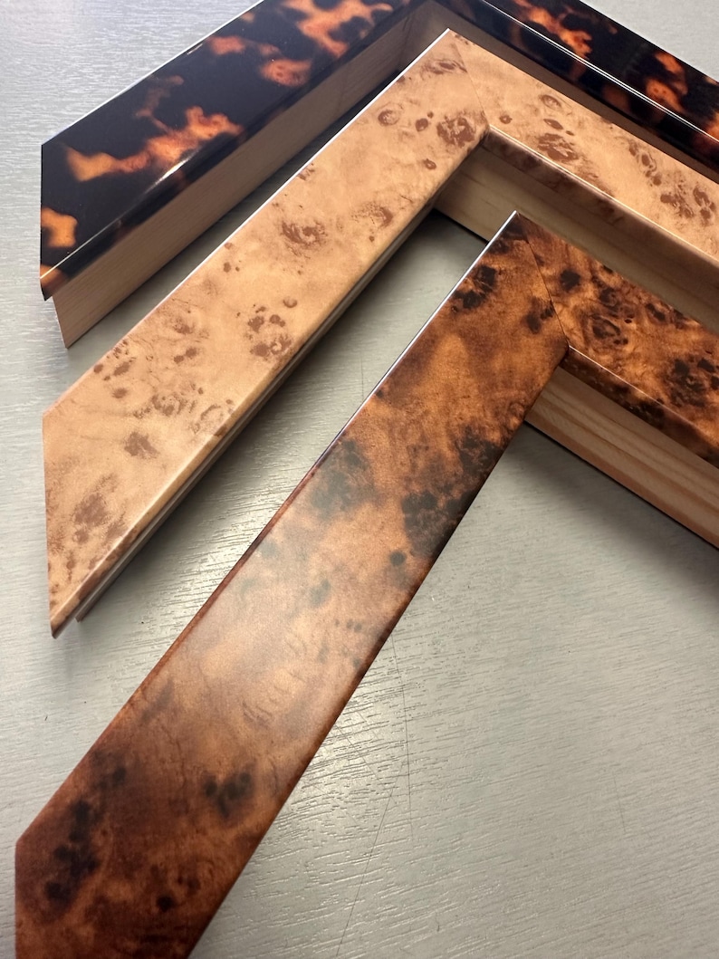 May include: Three L-shaped picture frame corner samples featuring distinct finishes. The top piece has a glossy tortoiseshell pattern, the middle piece displays a light tan burl wood grain, and the bottom piece shows a darker brown burl wood finish.