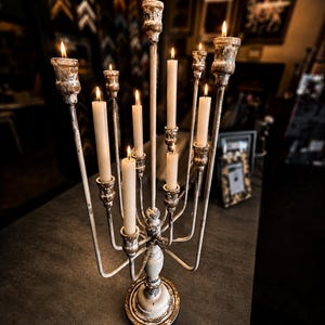 May include: A multi-armed metal candelabra with a distressed white and brown finish, featuring nine lit taper candles. The candelabra has a central stem and curved arms of varying heights, each topped with a candle cup holding a slender cream-coloured candle with a small flickering flame.