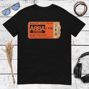 Könnte beinhalten: Ein schwarzes kurzärmeliges T-Shirt mit einem Grafikdruck eines orangefarbenen ABBA-Konzerttickets von 1979. Das Ticket enthält den Text ABBA IN CONCERT, Sportpaleis AHOY, 24 okt. 79 sowie Platzangaben für Sektion 3, Reihe K, Sitz 45.