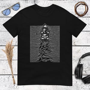 Puede incluir: Una camiseta negra de manga corta con un estampado gráfico blanco en el pecho. El diseño muestra un pequeño bote de remos con remos sobre una serie de líneas horizontales que forman un patrón ondulado similar a una señal de radio.