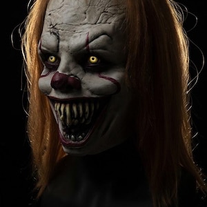 May include: A full-head clown mask featuring a pale, cracked skin texture, bright yellow eyes, and long orange hair. The mask displays a wide, menacing grin with sharp, pointed teeth, a red nose, and red facial markings extending from the eyes.