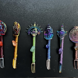 May include: Six decorative metal wands featuring iridescent wire-wrapped handles and various top ornaments including a pentagram, a hand with an eye, a sunburst, a shell, a dagger, and a circular sun emblem, each accented with a small raw gemstone.