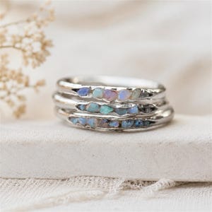 Puede incluir: Un conjunto de tres anillos de plata, cada uno con un canal engastado con pequeñas piedras de ópalo en bruto e iridiscentes. Las bandas de metal tienen un acabado liso y pulido, y los ópalos incrustados muestran tonos variados de azul, rosa y blanco.