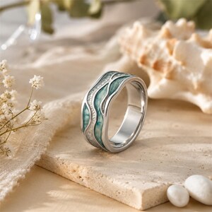 Puede incluir: Un anillo de tono plateado que presenta un canal ondulado y hundido relleno con un patrón jaspeado en color verde azulado y blanco. La banda tiene un acabado metálico liso y pulido con una forma orgánica curva y líneas fluidas.