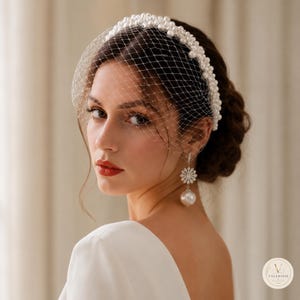 May include: A white headband adorned with numerous small white pearls, featuring an attached fine white netting veil that covers the upper face. The accessory is paired with a dangling earring consisting of a floral-shaped stud and a large pearl.