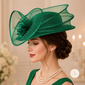 Op de afbeelding: Een smaragdgroene fascinatorhoed met een gestructureerd, breed ontwerp gemaakt van doorschijnende, gelaagde crinoline-stof. De hoed is versierd met een opvallend, geplooid bloemaccent aan de zijkant en bevat een klein logo met de tekst VALERISSE.
