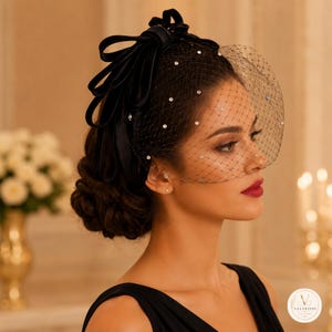 May include: A black fascinator headpiece featuring a large satin bow and a birdcage veil adorned with small, clear crystals. The veil covers the upper portion of the face, and the headpiece is secured with a structured black band.