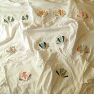 May include: A collection of white short-sleeved t-shirts, each featuring a central appliqué patch shaped like a seashell. The patches are constructed from various fabric segments in assorted colours and patterns, including polka dots and gingham.