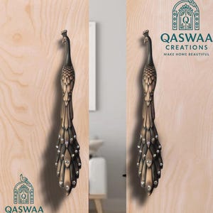 Op de afbeelding: Twee verticale deurklinken in de vorm van pauwen met een donkerbronzen afwerking en ingebedde heldere kristallen langs de staartveren. De klinken zijn gemonteerd op licht houten panelen met het logo van QASWAA CREATIONS erop.