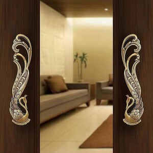 May include: Two matching brass-toned metal door handles mounted on dark wood doors. Each handle features an intricate, elongated peacock design with detailed feather patterns and a curved neck, positioned vertically on the door panels.