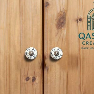 Può includere: Due pomelli rotondi in ceramica bianca con un motivo a foglie grigio scuro e centri metallici fissati su ante di armadi in legno marrone chiaro. Il logo QASWAA CREATIONS MAKE HOME BEAUTIFUL con un pavone è visibile in alto a destra.
