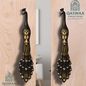 May include: A pair of dark bronze-toned metal door handles shaped like peacocks, featuring detailed feather textures and embedded clear crystals along the tail. The logo QASWAA CREATIONS MAKE HOME BEAUTIFUL is printed in the upper right and lower left corners.