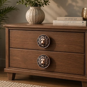 May include: A wooden nightstand with two drawers, each featuring a circular bronze-toned lion head knob. The top surface holds two stacked books with the text EAT DRINK NAP and LIVE BEAUTIFUL STYLE, alongside a metallic candle holder with cutouts.