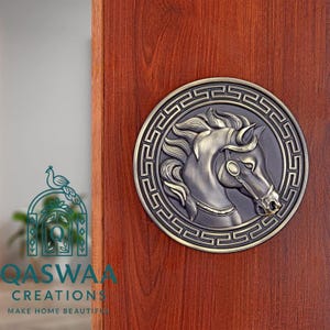 May include: A circular bronze-toned wall plaque featuring a relief sculpture of a horse head in profile, surrounded by a decorative Greek key border. The plaque is mounted on a vertical wooden surface with a visible grain pattern.