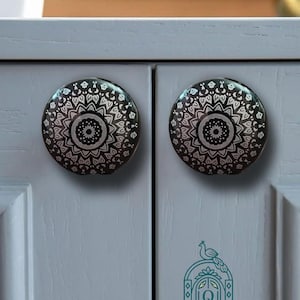 Puede incluir: Dos pomos de armario redondos con un patrón de mandala en blanco y negro. Los pomos están montados en puertas de armario de color gris claro. Debajo del pomo derecho, un logotipo verde azulado muestra un pavo real sobre un arco con el texto QASWAA CREATIONS MAKE HOME BEAUTIFUL.