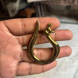 Könnte beinhalten: Ein poliertes, goldfarbenes Metallornament in Form eines stilisierten Pfaus mit einem geschwungenen, tropfenförmigen Körper. Das Design zeichnet sich durch eine glatte, reflektierende Oberfläche mit einem kleinen, detaillierten Kopf aus.
