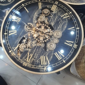 May include: A round wall clock with a black face featuring a gold-toned world map and exposed mechanical gears. The clock has large gold Roman numerals, gold hands, and gold-toned minute markers at 15, 30, 45, and 60, all encased in a gold frame.