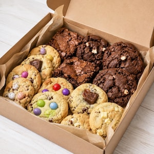 May include: A cardboard box filled with an assortment of baked goods, including chocolate brownies, dark chocolate chip cookies, and light-coloured cookies topped with colourful candy-coated chocolate eggs and various chocolate chunks.