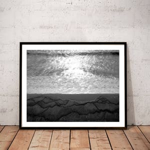 May include: Black and white photograph of a beach scene with waves crashing on the shore. The water is reflecting the sunlight, creating a shimmering effect. The sand is textured and appears to be wet.