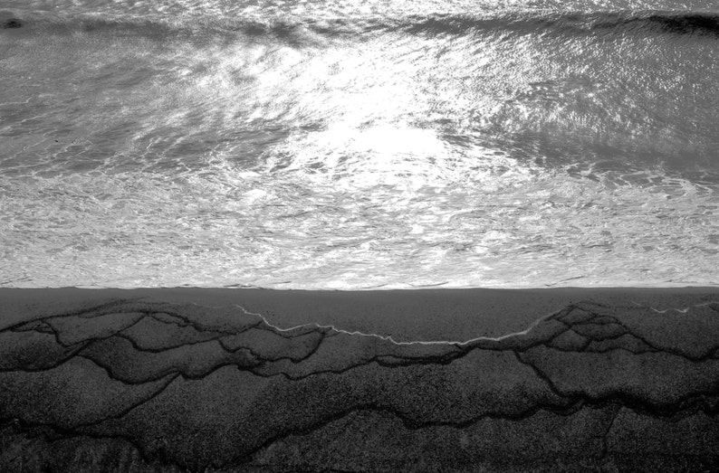 May include: A black and white photo of a beach with waves crashing on the shore. The sand is wet and has a pattern of lines.
