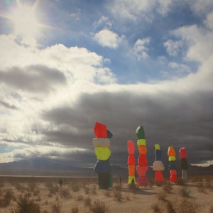 May include: Seven colorful, stacked, geometric sculptures in a desert landscape. The sculptures are made of painted concrete and are arranged in a row. The sky is cloudy and the sun is shining brightly.