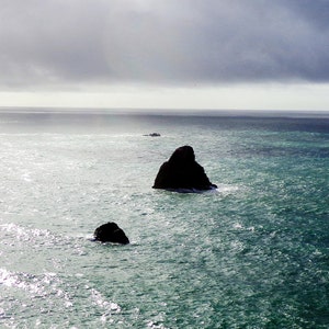 K&ouml;nnte beinhalten: Drei dunkelgraue Felsen ragen aus dem Ozeanwasser heraus. Das Wasser hat eine hellblaugr&uuml;ne Farbe und der Himmel ist dunkelgrau mit einem Hauch von Licht in der Ferne.