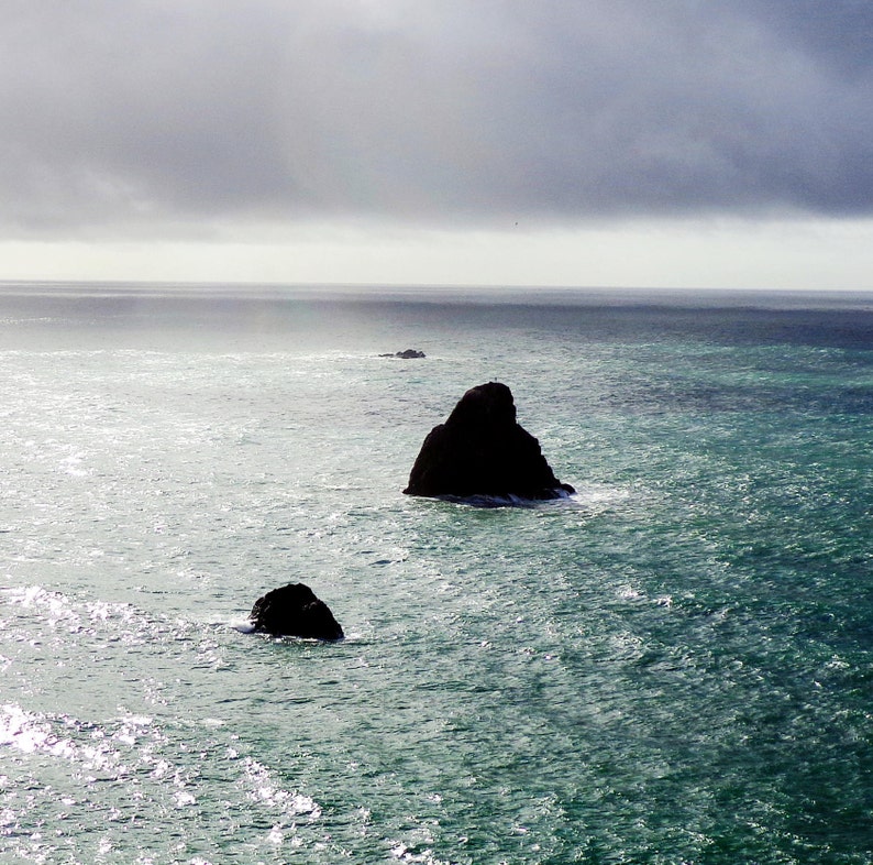 K&ouml;nnte beinhalten: Drei dunkelgraue Felsen ragen aus dem Ozeanwasser heraus. Das Wasser hat eine hellblaugr&uuml;ne Farbe und der Himmel ist dunkelgrau mit einem Hauch von Licht in der Ferne.