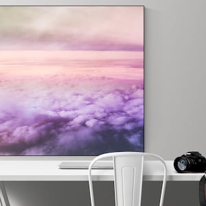May include: A framed photograph of a pink and purple sky with clouds. The photograph is hanging on a white wall above a white desk with a white chair, a camera, and a mug.