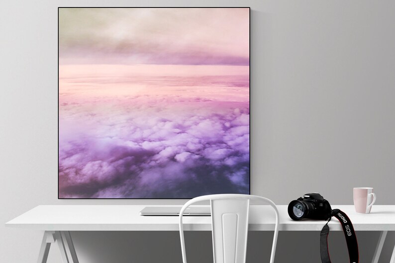 May include: A framed photograph of a pink and purple sky with clouds. The photograph is hanging on a white wall above a white desk with a white chair, a camera, and a mug.