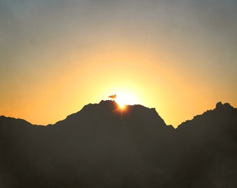 以下が含まれることがあります： 山脈のシルエットが、その背後に昇る太陽によって、空に暖かく金色に輝いています。