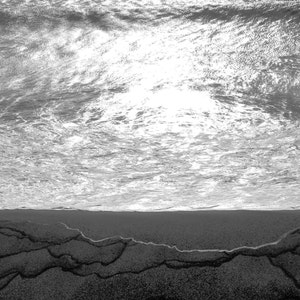 May include: A black and white photo of a beach with the ocean waves crashing on the shore. The sand is wet and there are ripples in the sand from the receding waves.