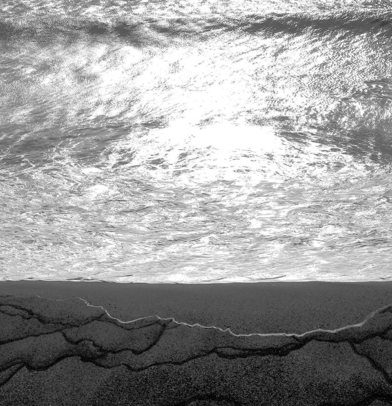 May include: A black and white photo of a beach with the ocean waves crashing on the shore. The sand is wet and there are ripples in the sand from the receding waves.