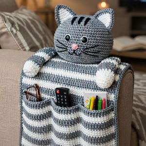 Könnte beinhalten: Ein grau-weiß gestreifter Häkel-Armlehnen-Organizer mit einem Katzenkopf oben, weißen Innenohren, schwarzen Schnurrhaaren und einer rosa Nase. Der Organizer hat drei Taschen, die eine Brille, eine Fernbedienung und mehrere Stifte enthalten.