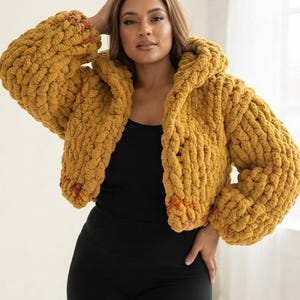 May include: A mustard yellow chunky knit hooded cardigan featuring an oversized, thick yarn texture with long sleeves and an open front design. The garment is worn over a simple black tank top and black leggings, showcasing a cropped silhouette.