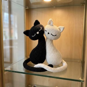 Puede incluir: Dos figuras de gato tejidas a ganchillo, una negra y otra color crema, sentadas una al lado de la otra con sus largas colas entrelazadas. Cada gato presenta ojos cerrados bordados y una nariz pequeña con textura uniforme.