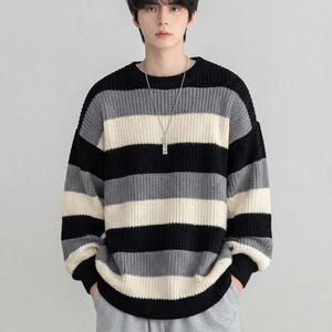 May include: A long-sleeved crew neck sweater featuring horizontal stripes in black, charcoal gray, and off-white. The knit garment has a ribbed collar, cuffs, and hem, with a thick, textured weave pattern throughout the body and sleeves.