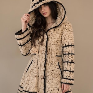 May include: A beige crochet hooded coat featuring black stitched detailing on the sleeves and hem, a large hood with black accents, and a front button closure with round wooden buttons. The garment has a textured knit pattern throughout.