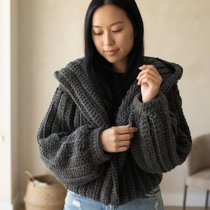 May include: A dark charcoal gray chunky knit hooded cardigan featuring a thick ribbed texture, long sleeves with dropped shoulders, and an open front design. The garment is crafted from a thick yarn with a visible vertical stitch pattern throughout.
