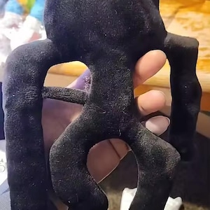 May include: A black plush toy featuring a flat, rectangular head, two long arms extending from the sides, and two shorter, curved legs at the bottom. The soft, fabric-covered figure has a simple, abstract silhouette with a smooth, dark texture.