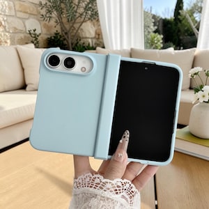 May include: A light blue foldable smartphone case with a matte finish, featuring a dedicated cutout for the rear camera module and a hinged section that protects the spine of the device while leaving the inner screen exposed for full functionality.