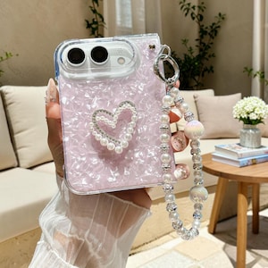 May include: A light pink, pearlescent mobile phone case featuring a heart-shaped embellishment made of white faux pearls and clear crystals. The case includes a matching wrist strap composed of white faux pearls and silver-toned beads.